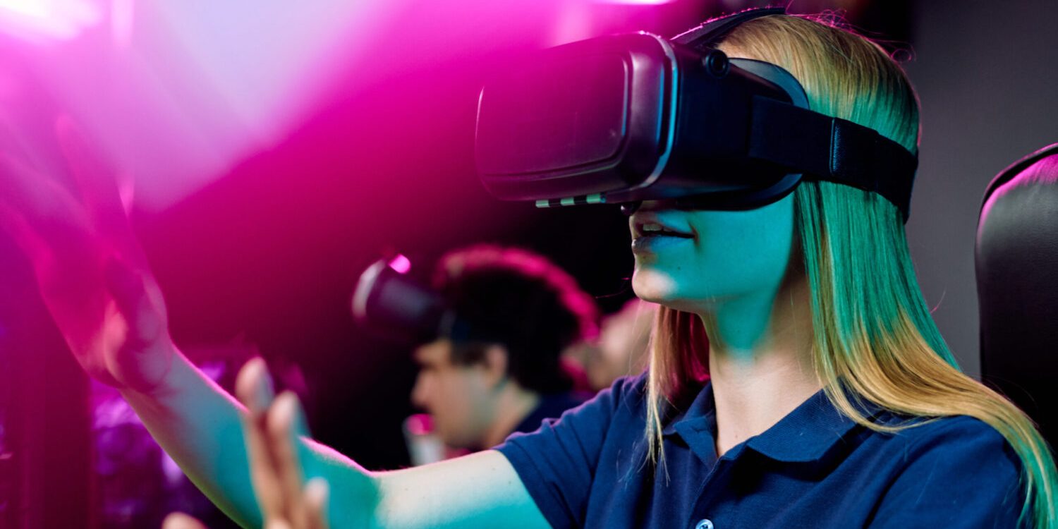 Blond teenage girl in vr goggle touching virtual display while taking part in cybersports video game on background of her friends